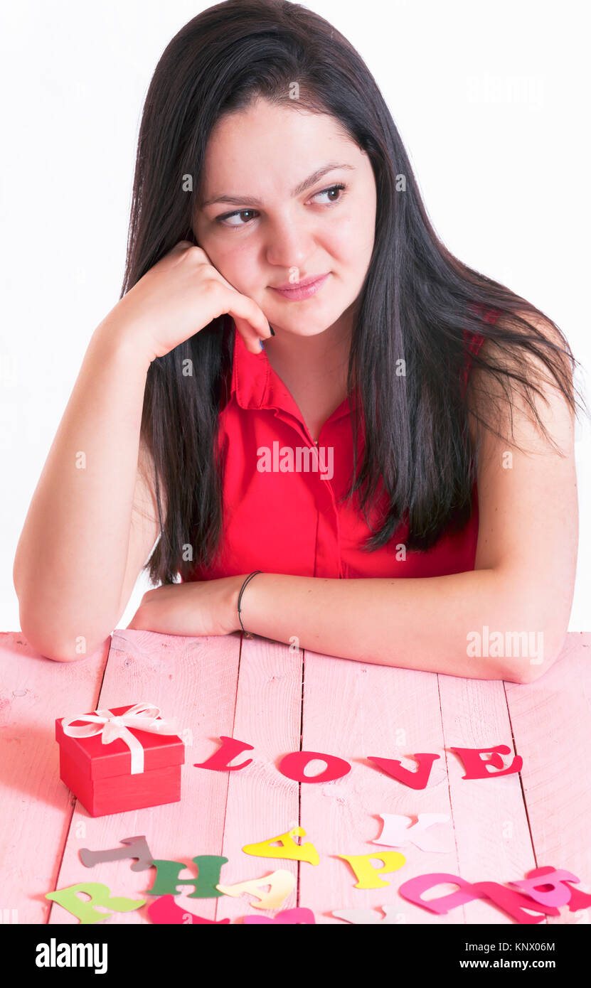 Attractive brunette woman thinking while sitting at a pink table, with a red gift and the word love spelled from red paper letters in front of her. Stock Photo