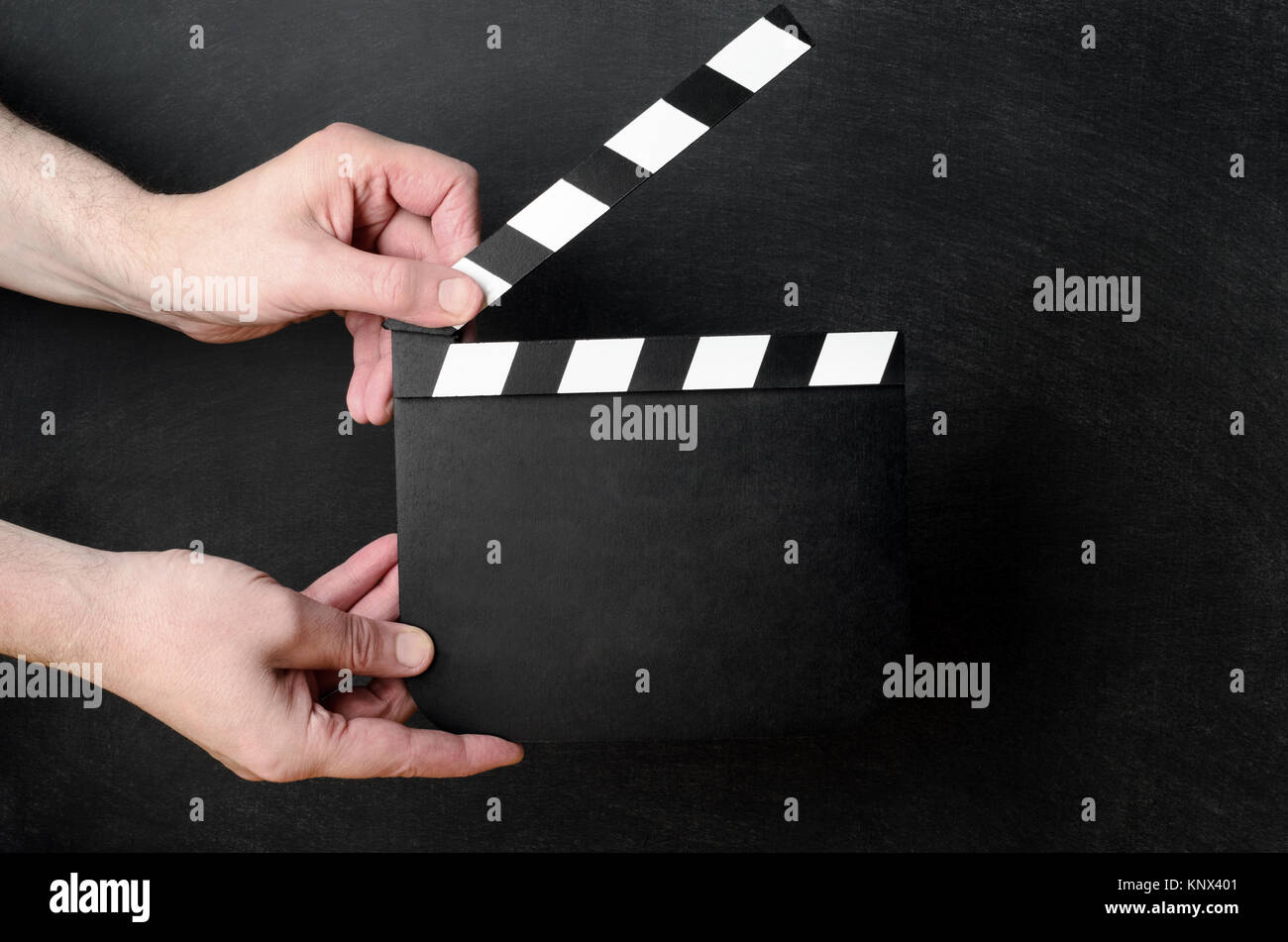 A blank clapperboard held open as if about to snap shut between the hands of a male operative with black chalkboard background. Stock Photo