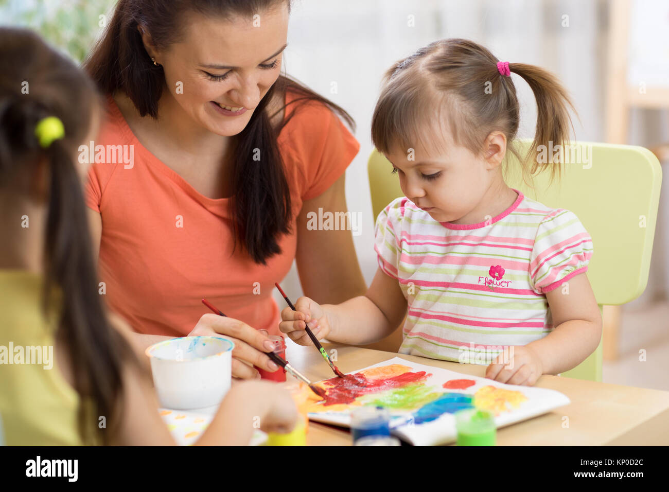 Teacher and little girls are painting in daycare center. Woman and children have a fun pastime. Stock Photo