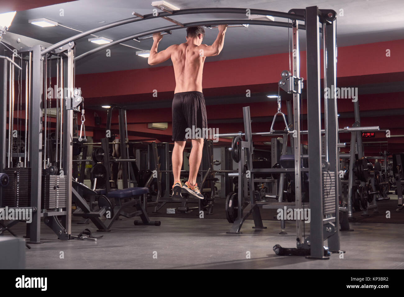 one young man, Multi-Station Gym Machine, back exercise, rear view, dark gym indoors, full lenght body shot. Stock Photo