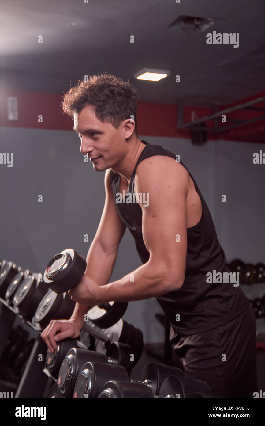 one young man looking smirking, picking dumbbells, dark gym indoors. Stock Photo