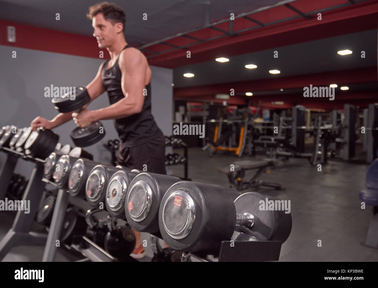 one young man looking smirking, picking dumbbells, man out of focus, focus on dumbbells rack, dark gym indoors. Stock Photo