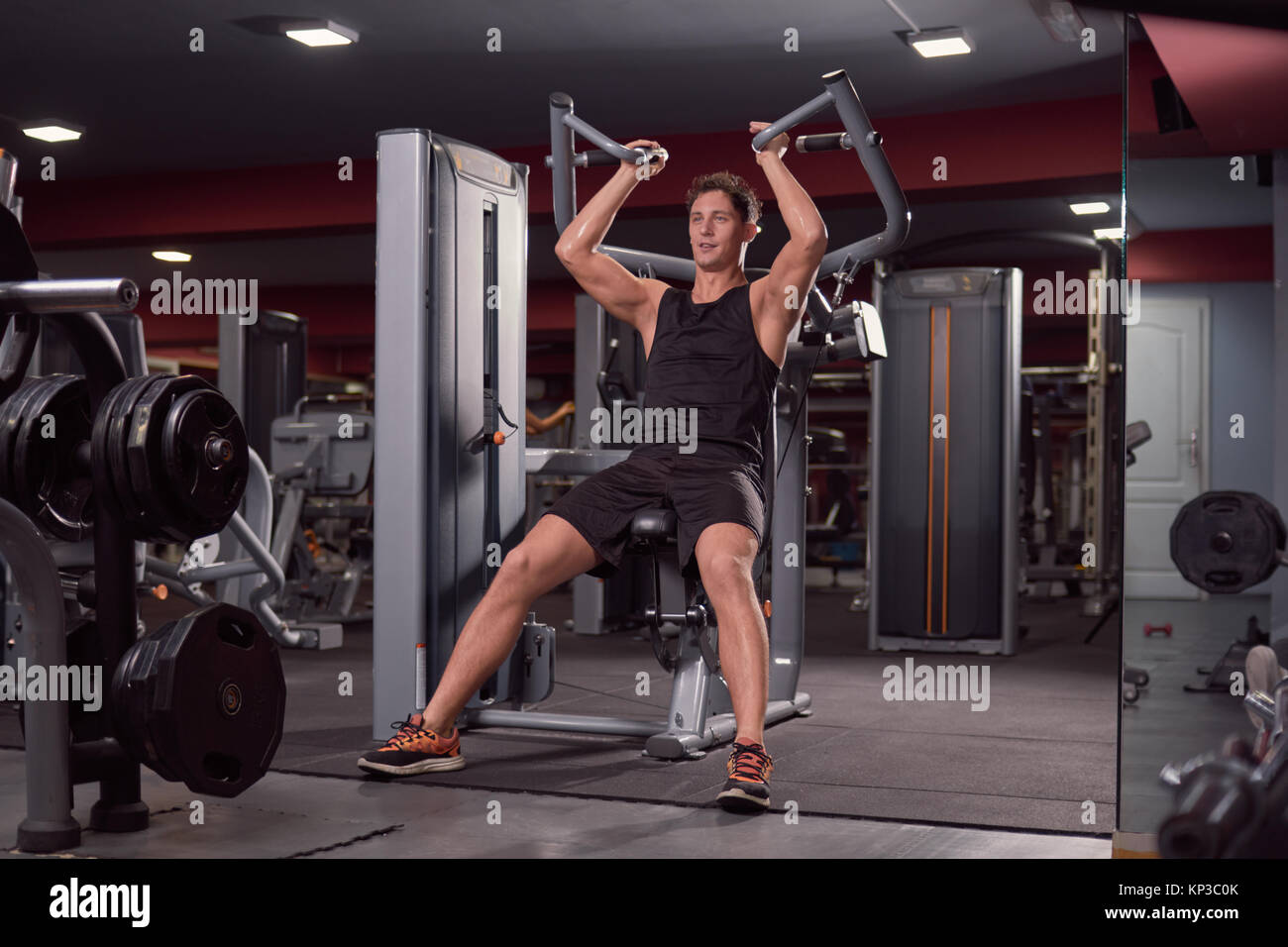 one young man, fitness machine, seated back lat pull down, dark gym indoors. Stock Photo
