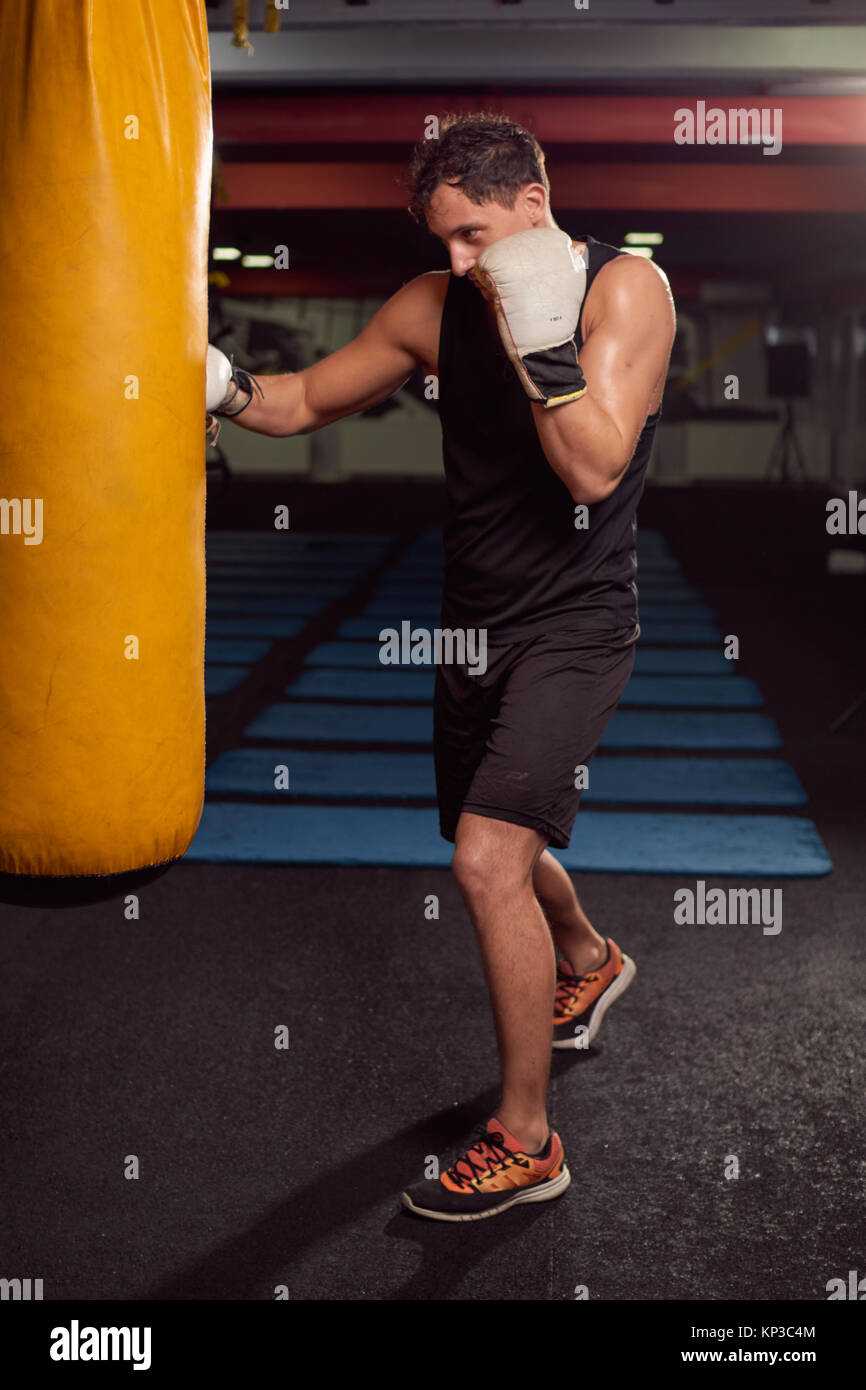 one young man, boxer hand hitting punching bag, practicing indoors gym room, wearing boxing gloves, full lenght body shot. Stock Photo