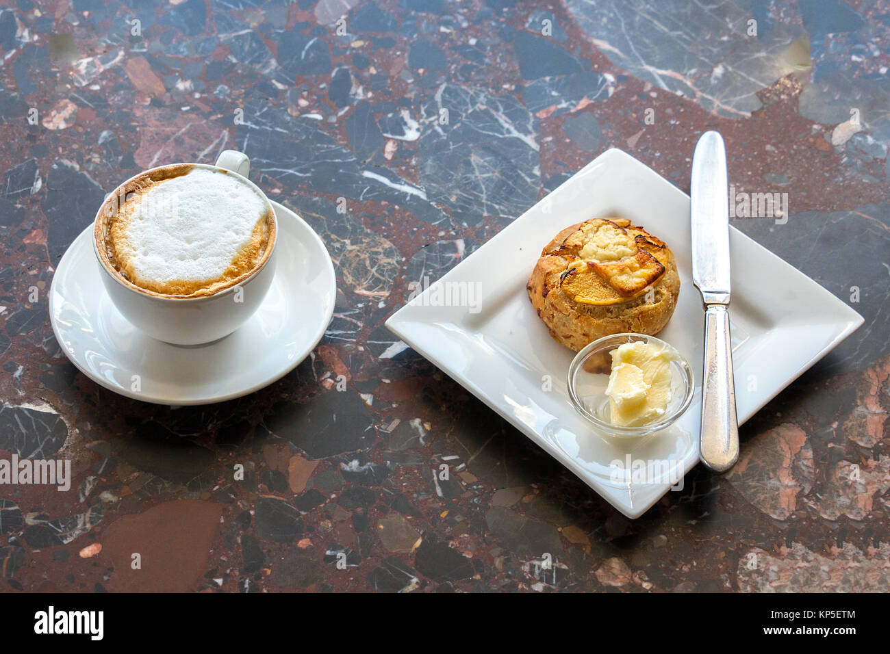 Hight view of a white cup of cappuccino with a scone with butter on a plate with a knife on a table, indoors. Stock Photo