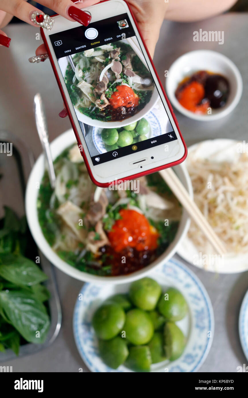 Bowl of Vietnamese noodle soup known as Pho. Woman making picture with smartphone of her meal. Ho Chi Minh City. Vietnam. Stock Photo