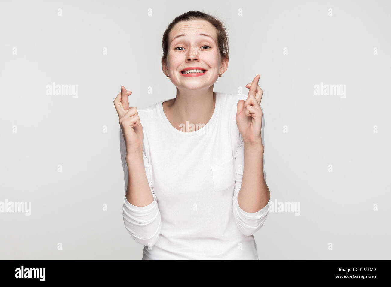 Young adult beautiful girl toothy smiling and looking at camera, crossed fingers. Studio shot,isolated on gray background Stock Photo