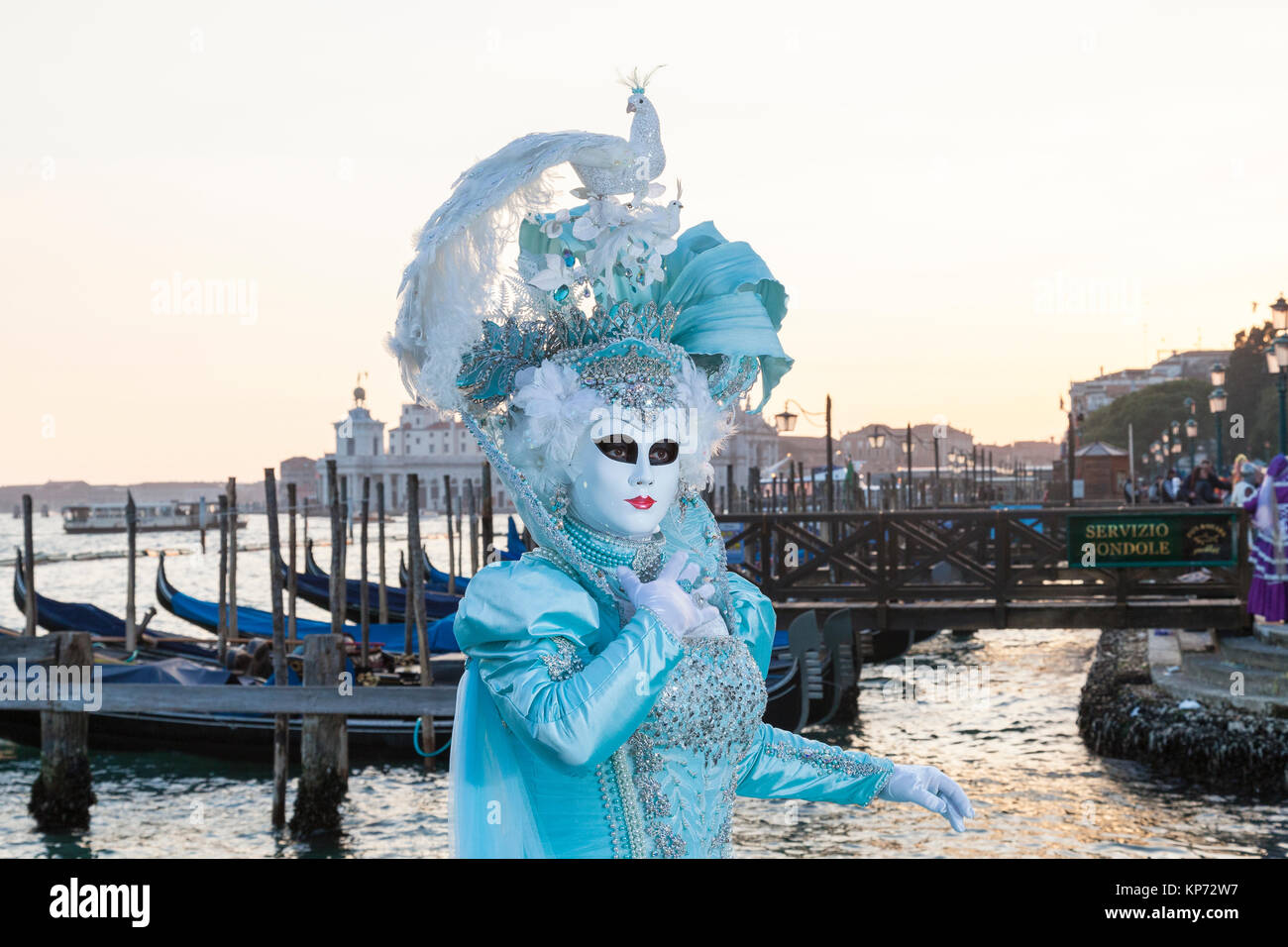 Venice Carnival, Venice, Italy. Woman in a peacock headdress posing at the lagoon at sunset with gondolas Stock Photo