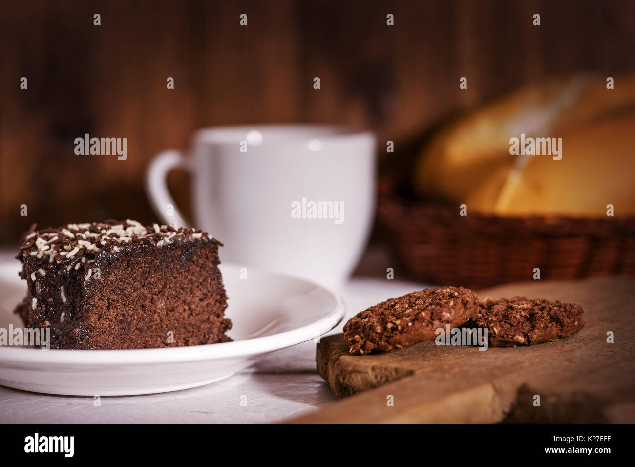 Chocolate cake, cookies, coffee porcelain cup and bread loafs on top of a wooden white table top and wood background Stock Photo