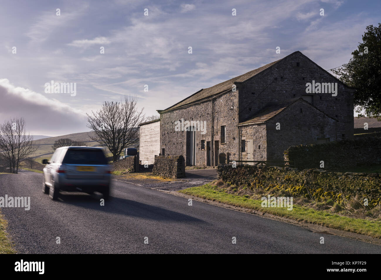 Car (Land Rover Freelander) travelling past stone, roadside barn & farm buildings on country road - Barden, Yorkshire Dales, England, UK. Stock Photo