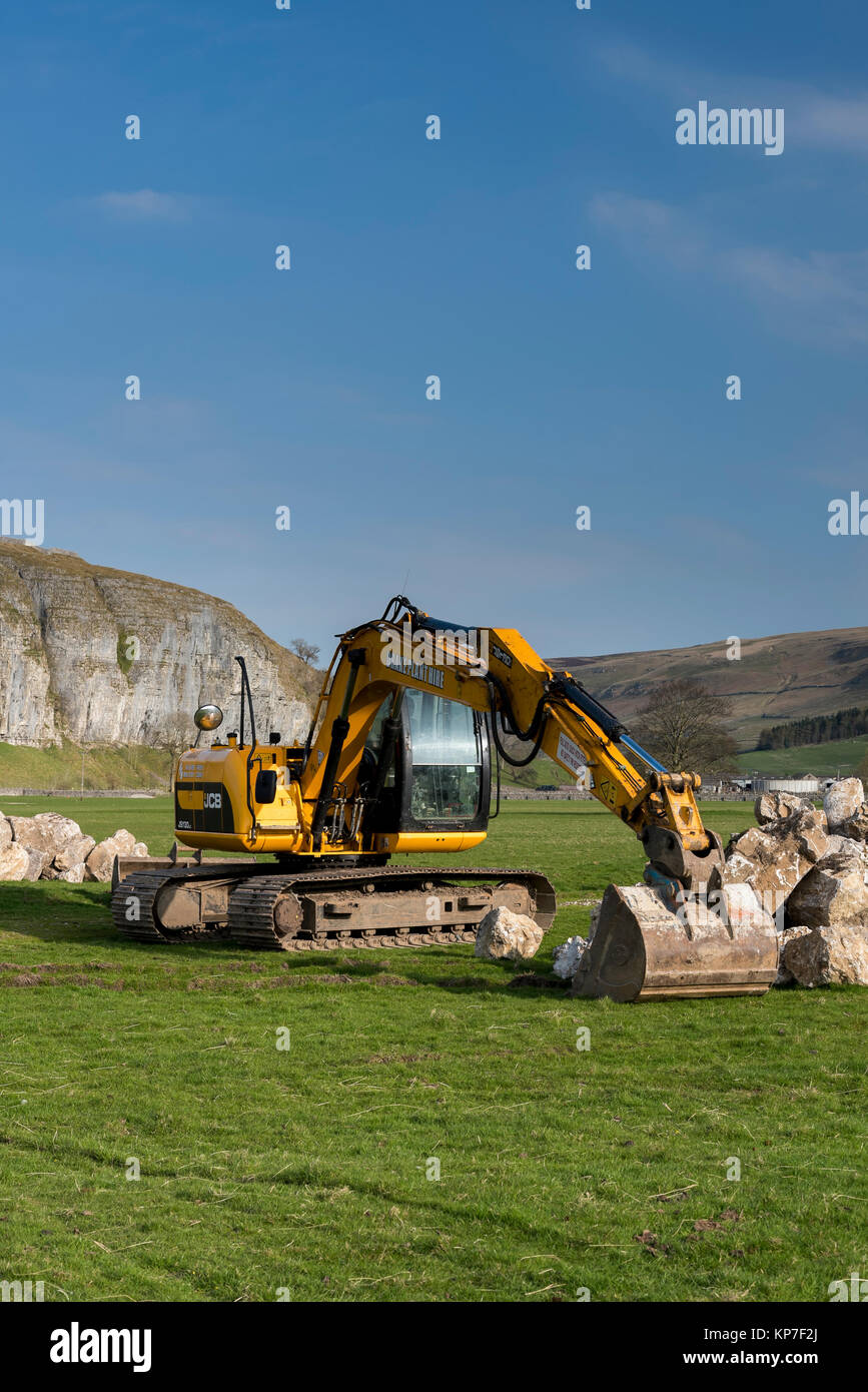 Bright yellow JCB JS130LC tracked excavator standing in countryside farm field, ready to lift heavy boulders - Kilnsey, Yorkshire Dales, England, UK. Stock Photo