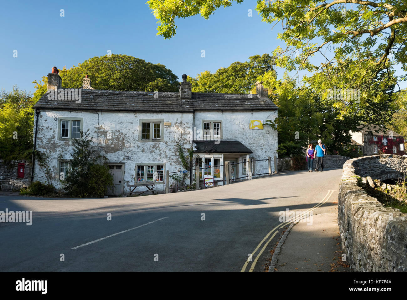 On sunny day, couple are walking across stone bridge in Malham centre,  by old village shop with peeling white paint - Yorkshire Dales, England, UK. Stock Photo