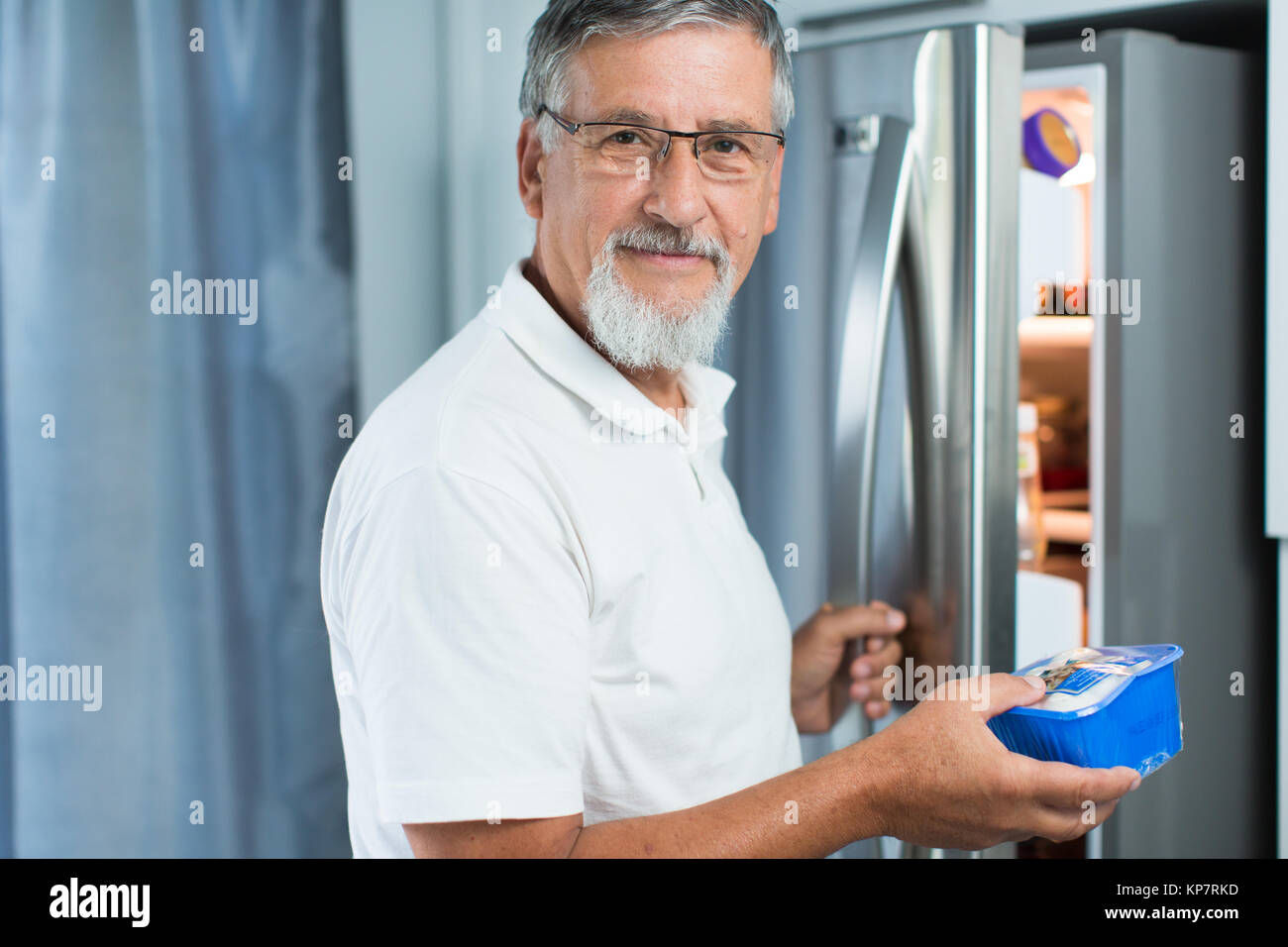 Is this still fine? Senior man in his kitchen by the fridge, looking at the expiry date of a product she took from her fridge - Stock Photo