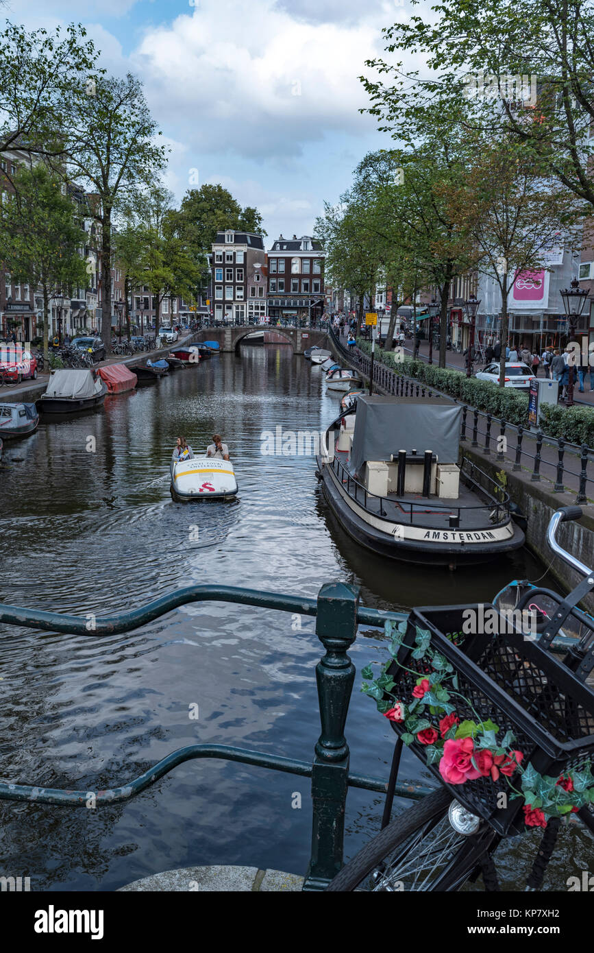 Moored Boats line a Canal in Amsterdam with Bicycle and Flowers in Foreground Stock Photo