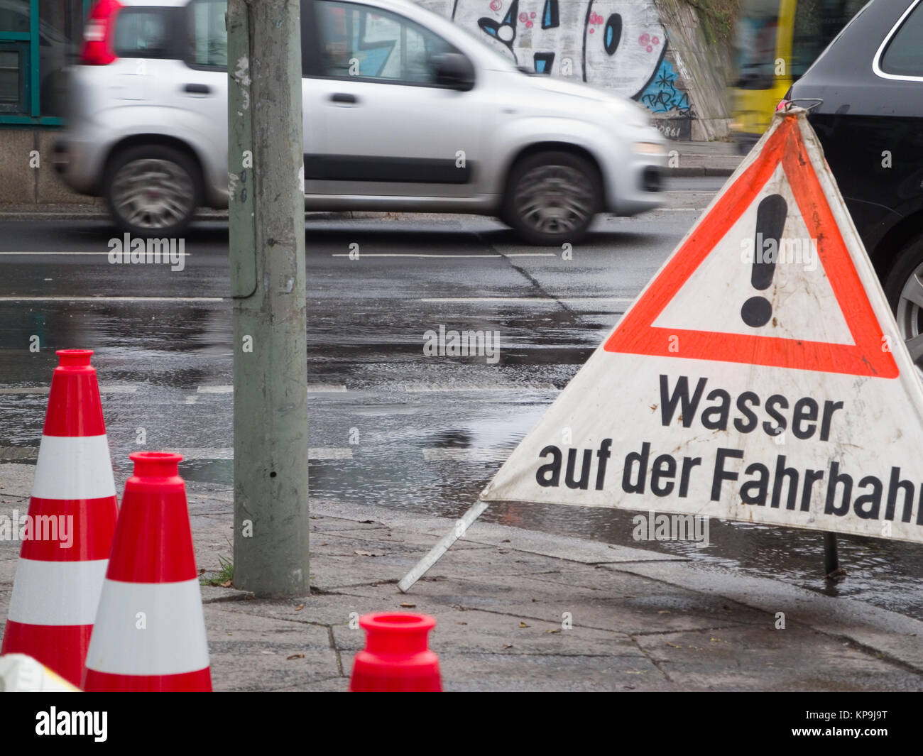 Detail-Ansicht einer regennassen Straßenkreuzung mit Personsnkraftfahrzeug in Berlin mit aufgestellten Absperrungen und der Aufschrift 'Wasser auf der Fahrbahn' Stock Photo