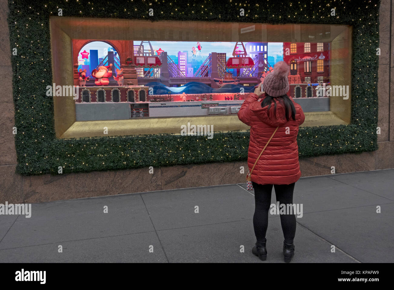 A woman in an orange down coat taking photos of a  Macy's Christmas window at Herald Square in Manhattan, New York City. Stock Photo