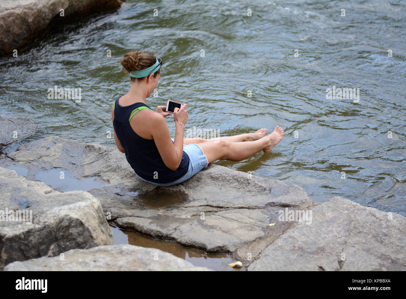 Woman checking her phone while relaxing and cooling off while dipping her legs in water of creek Stock Photo