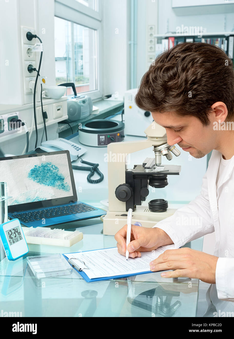 Young male scientist or tech makes notes in histopathology laboratory with light microscope and a laptop. Shallow DOF, focus on the right hand, Stock Photo
