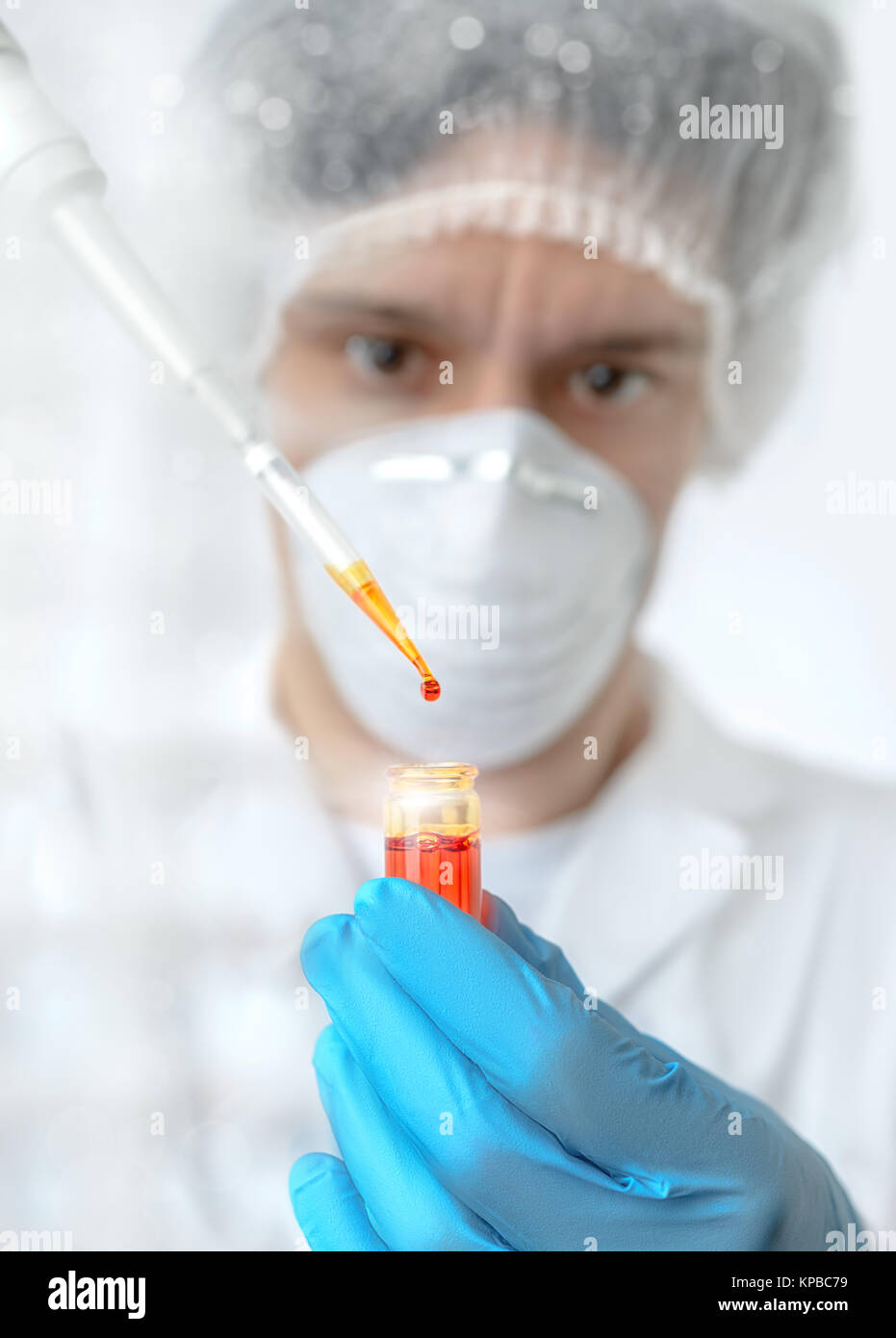 Keen scientist with brown eyes in protective wear pipettes orange sample, Shallow DOF, focus on the drop Stock Photo
