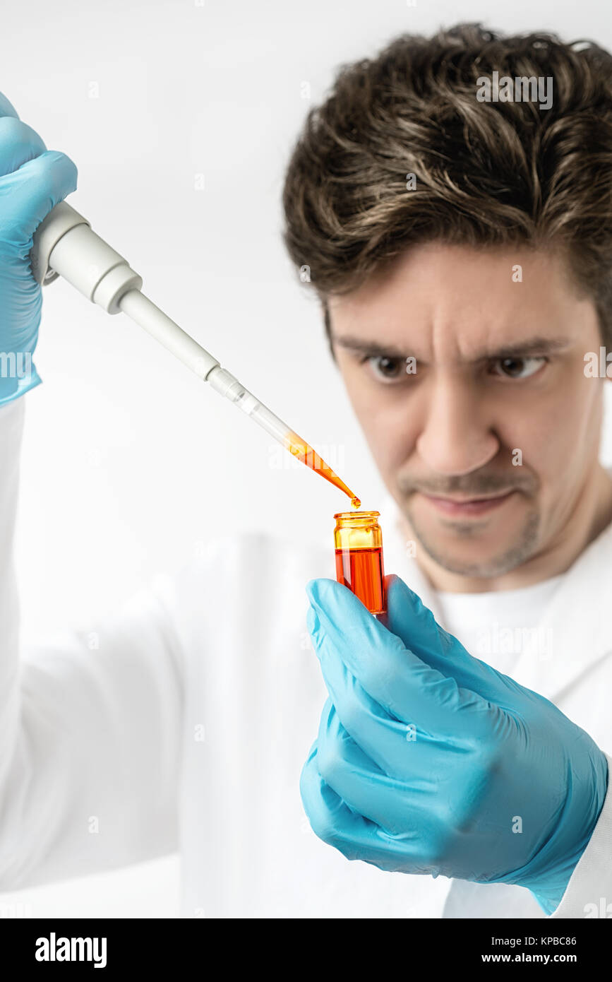 Keen scientist with brown eyes in protective wear pipettes orange sample, Shallow DOF, focus on the pipette, drop, tube and gloved hand. This image is Stock Photo