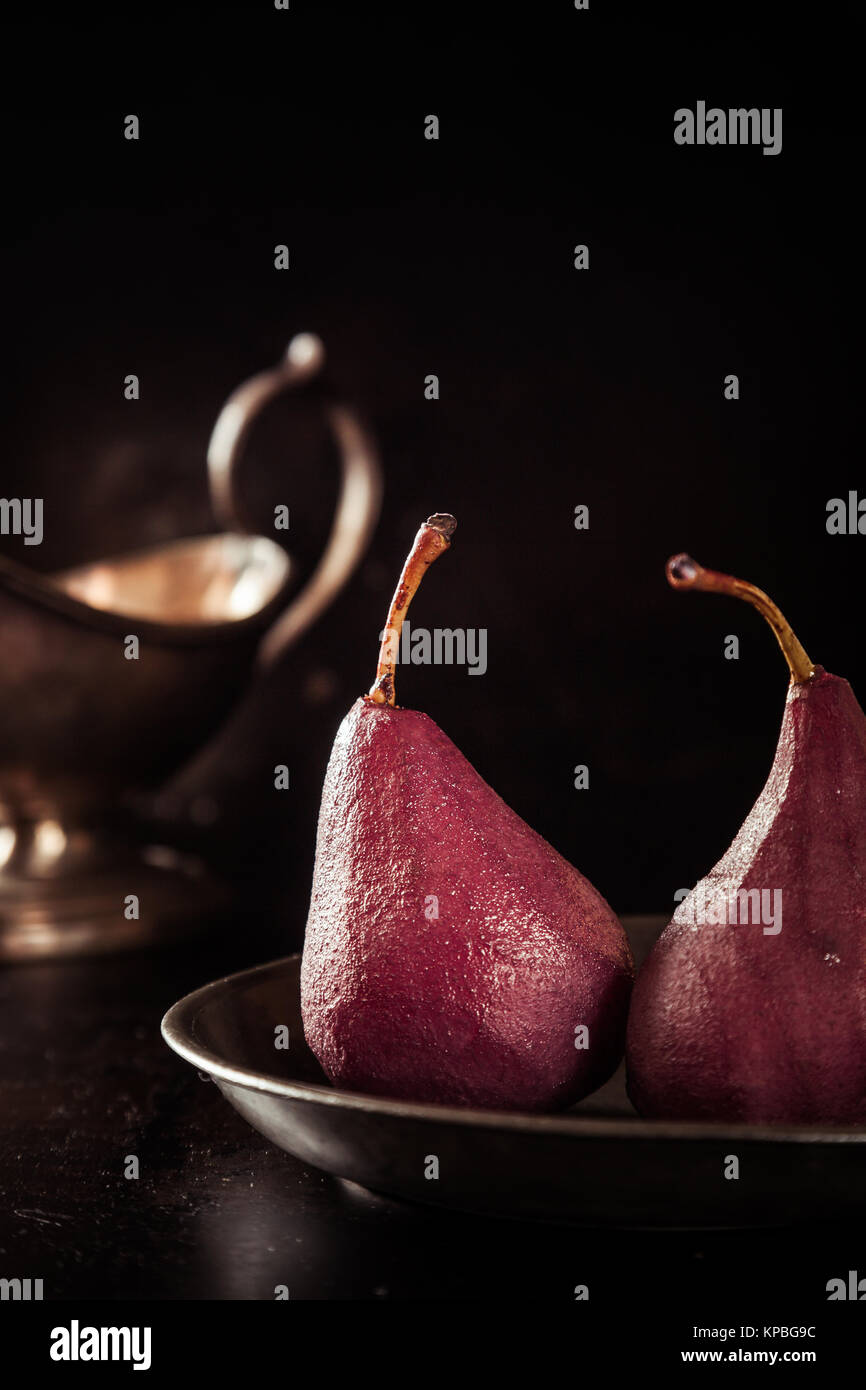 Glazed fresh whole pears steeped in red wine served for a speciality dessert with a silver sauce boat alongside in a dark shadowed environment Stock Photo