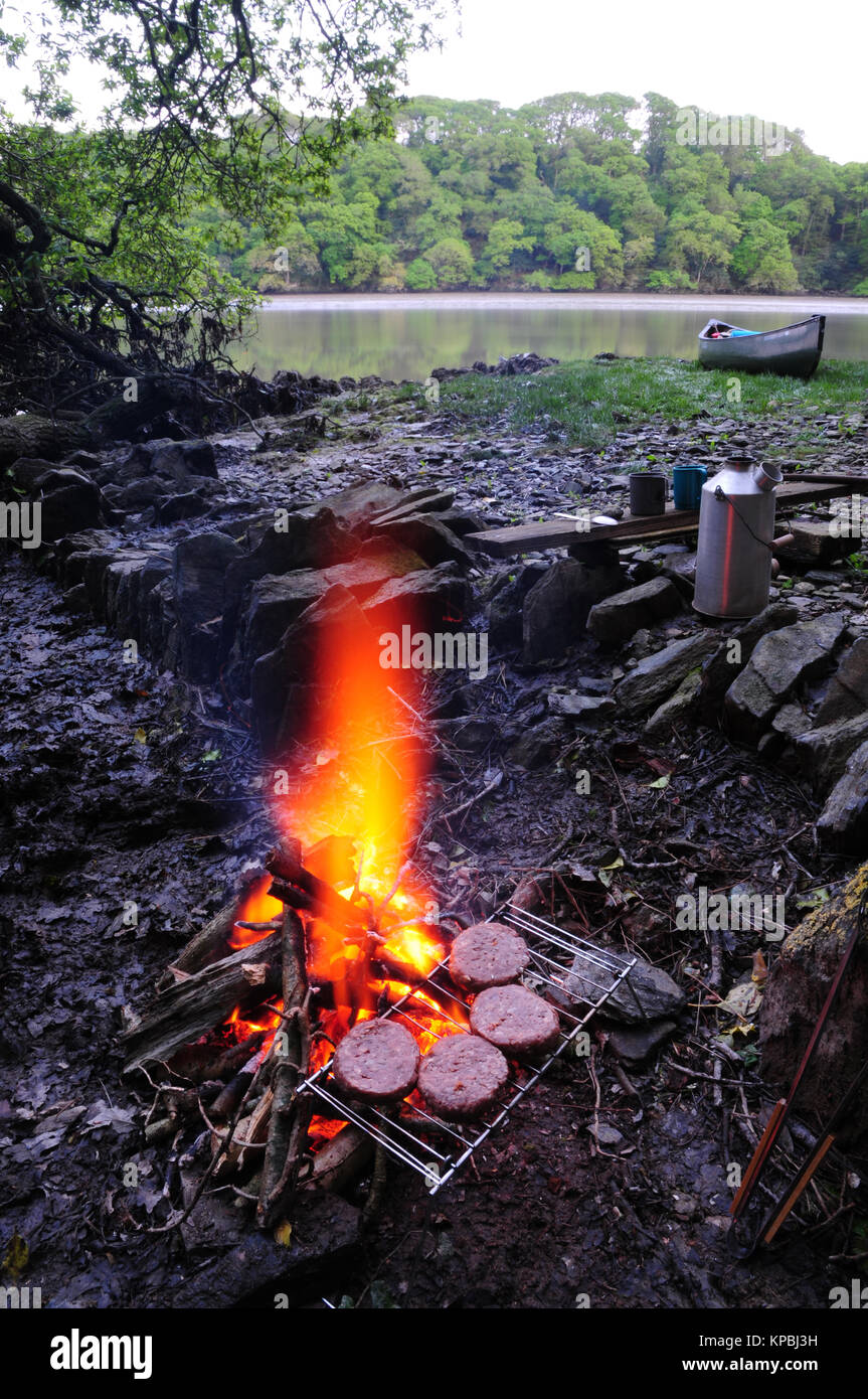 Cooking beef burgers on an open fire while canoe camping. In the background is a Kelly kettle