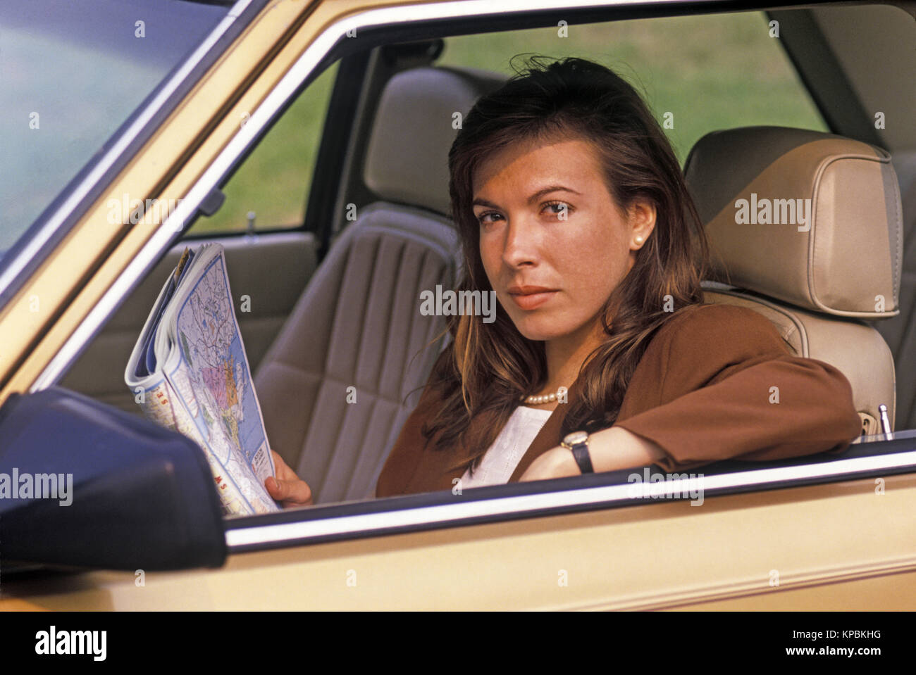 1988 HISTORICAL OFFICE WORKER SITTING IN DRIVERS SEAT OF LEFT HAND DRIVE AUTOMOBILE (©RENAULT S A  / AMC 1983) HOLDING US ROAD ATLAS (©RAND MCNALLY 1985) Stock Photo