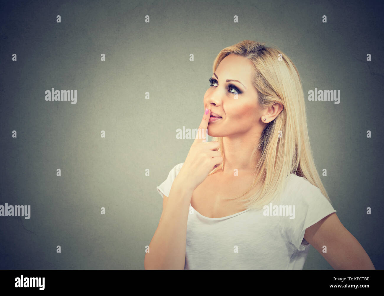 Young blonde touching lips while thinking on something happily and looking away on gray background Stock Photo