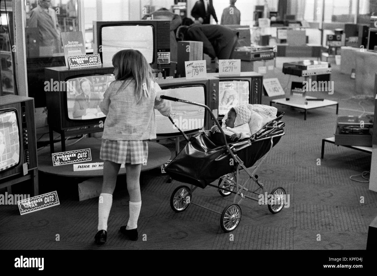 Multi ethnic Britain 1970s, white girls pushing black British baby in a pram around a department store London 1972. Uk department store selling televisions Signs say 'Purchase Tax Cuts'. Dickie Davies ITV World of Sport TV presenter is on the telly. HOMER SYKES Stock Photo
