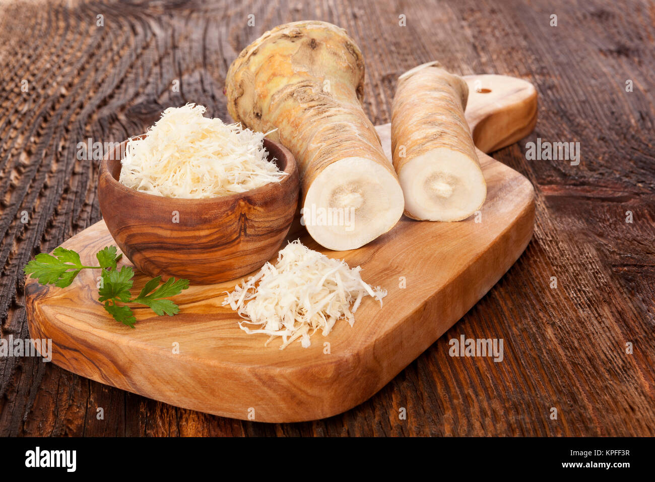 Fresh and grated horseradish in wooden bowl with parsley on wooden cutting board. Horse-radish root. Stock Photo