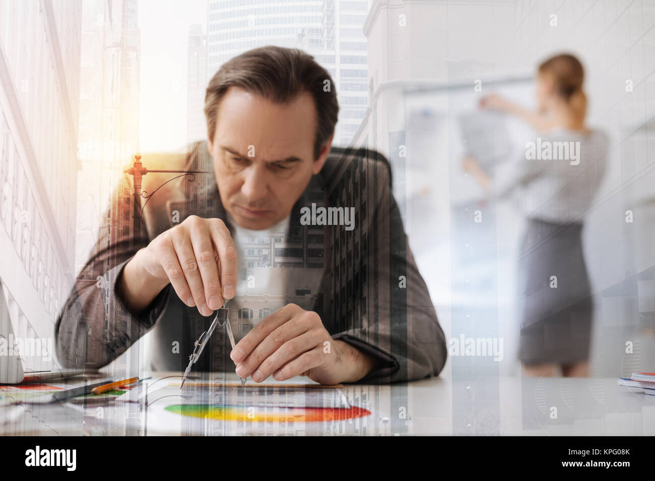Stick to accuracy. Waist up of a serious senior engineer sitting at the table and using compasses while his female colleague standing in the backgroun Stock Photo