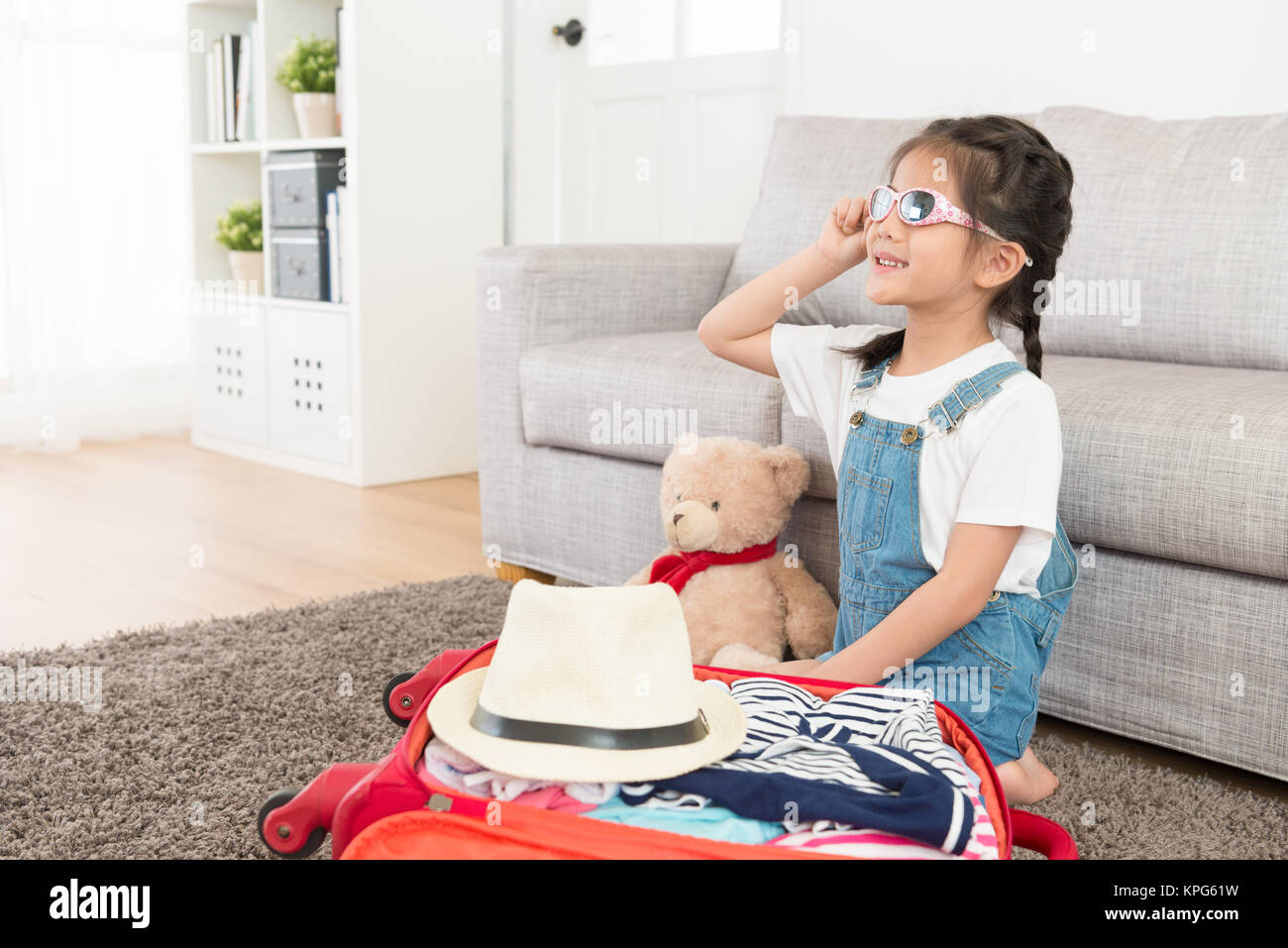 smiling happy girl children wearing sunglasses sitting on living room floor with teddy bear and finished packing luggage suitcase making daydreaming g Stock Photo