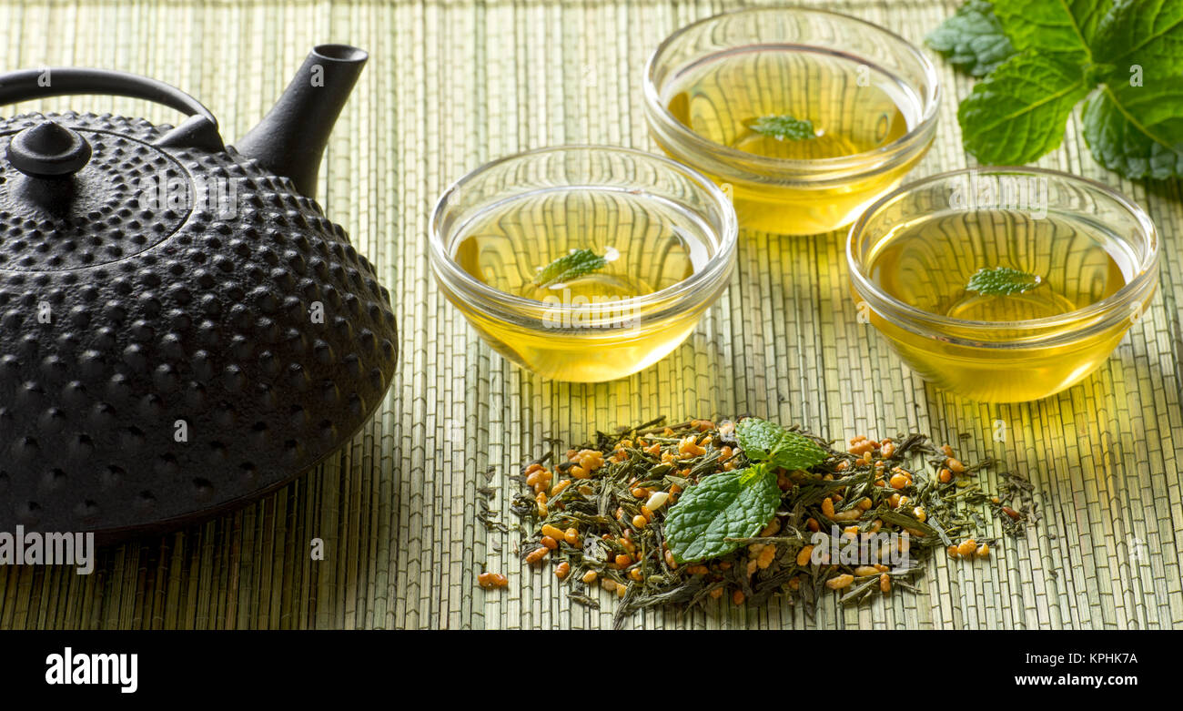 Japanese Fresh Organic Green Tea and tea leaves with clear drinking glasses and metal teapot sitting on bamboo mat background Stock Photo