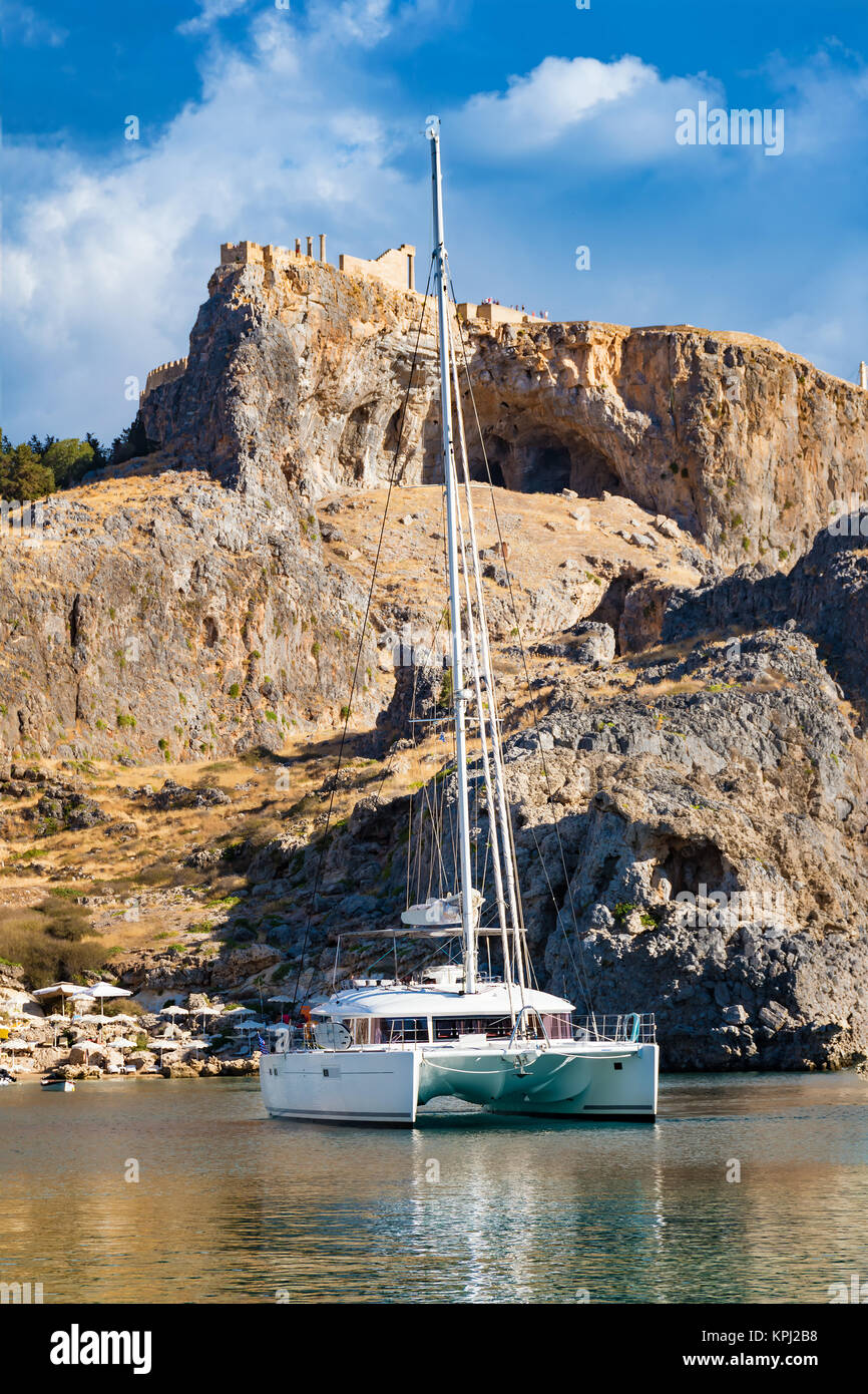 Catamaran in St. Paul´s bay, cloudy blue sky, Lindos acropolis in background (Rhodes, Greece) Stock Photo