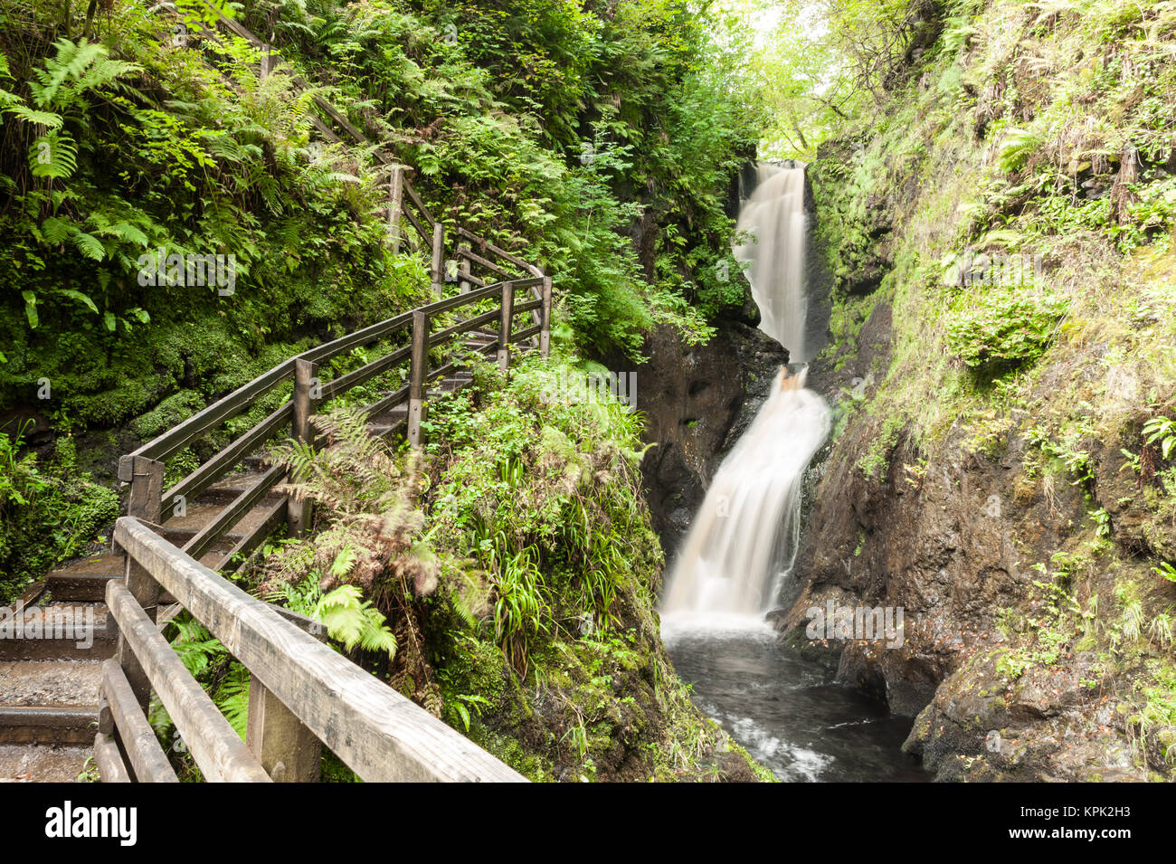Footpath outdoor trail track with wooden fence next to a small fast flowing waterfall in a gorge in the forest. Glenariff Forest Park, Northern Irelan Stock Photo