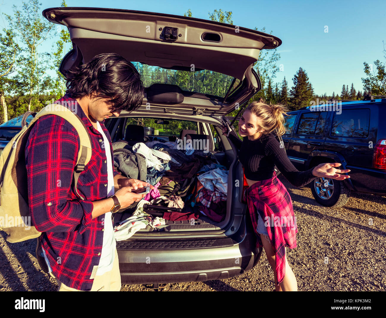 Young couple standing at the back of their packed vehicle with back door open and the young man in using his cell phone while the young woman waits... Stock Photo
