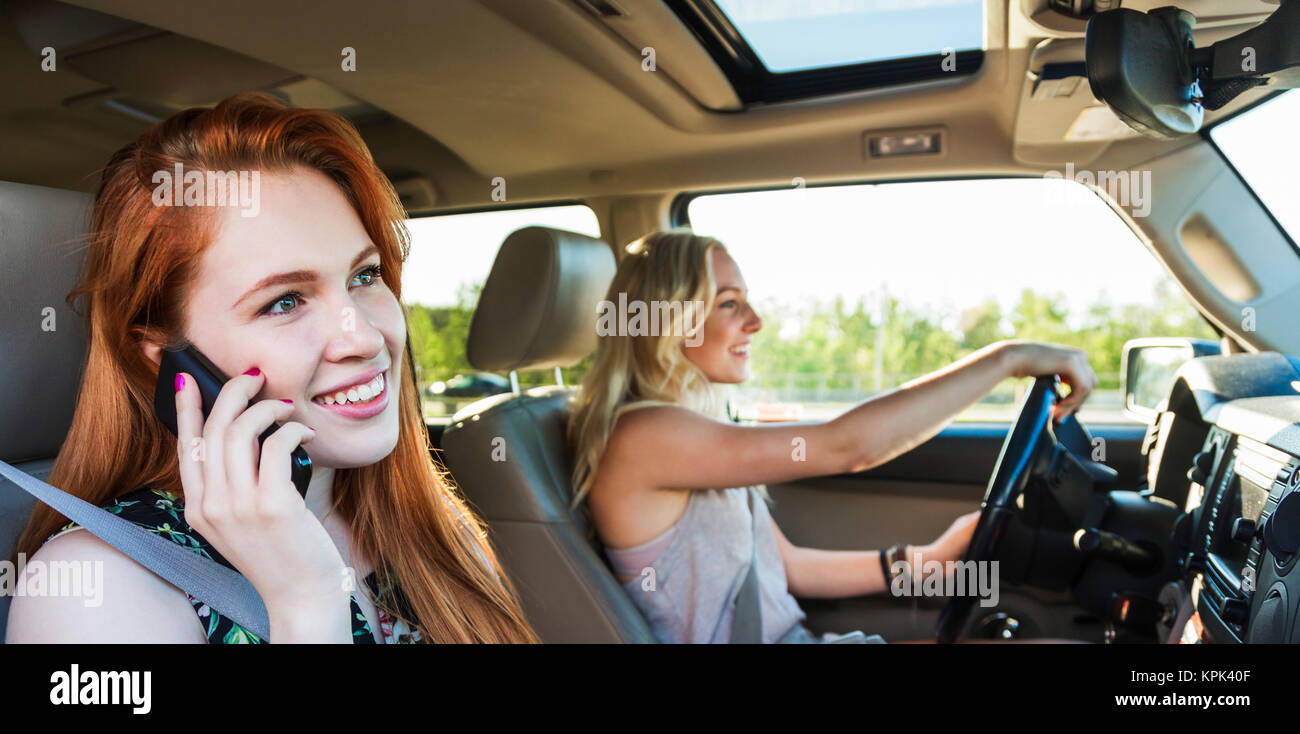 Two young women driving in a car on a road trip while one talks on a smart phone; Edmonton, Alberta, Canada Stock Photo
