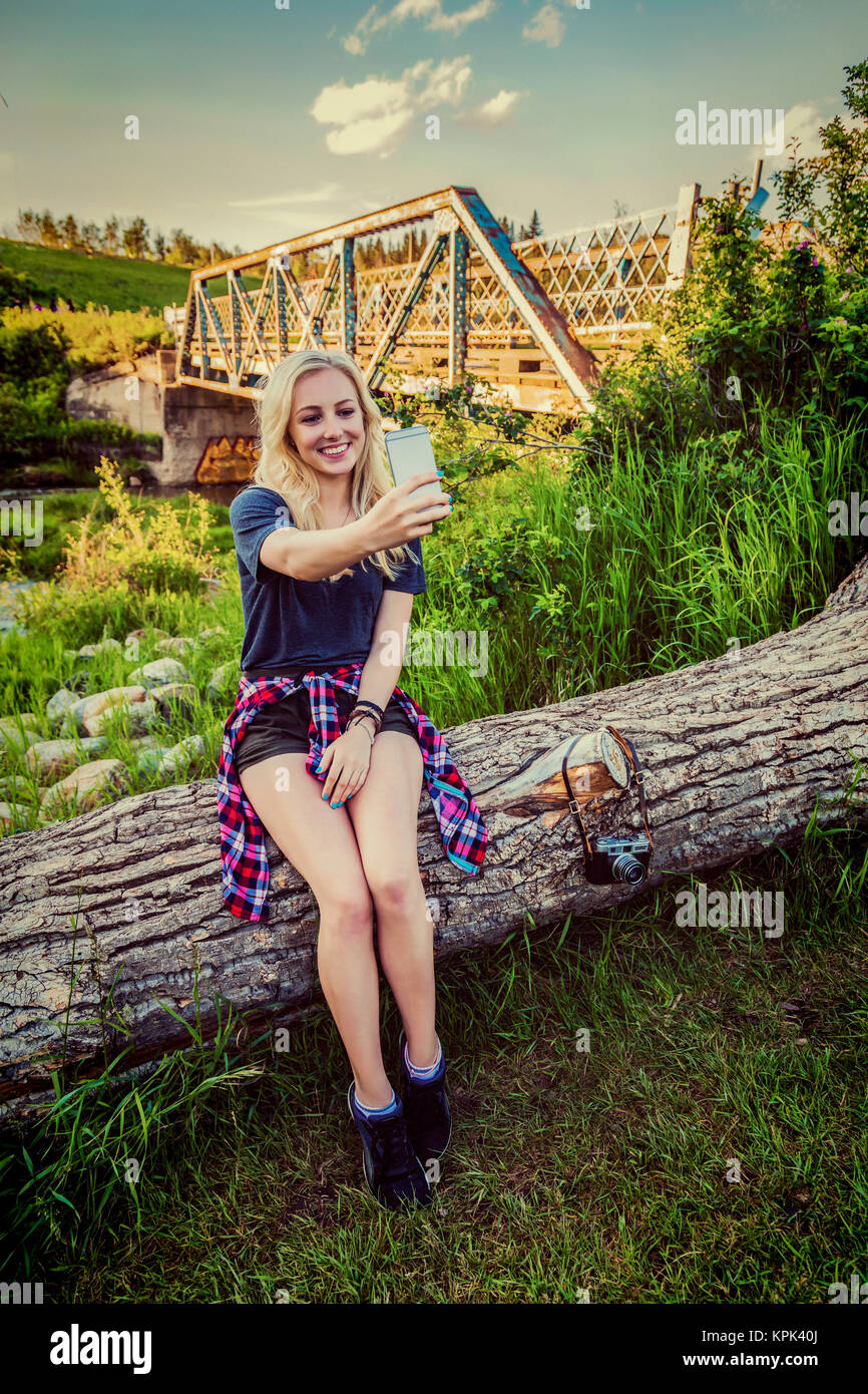 A young woman sits on a log in a park taking a self-portrait with her cell phone with a bridge and river in the background; Edmonton, Alberta, Canada Stock Photo