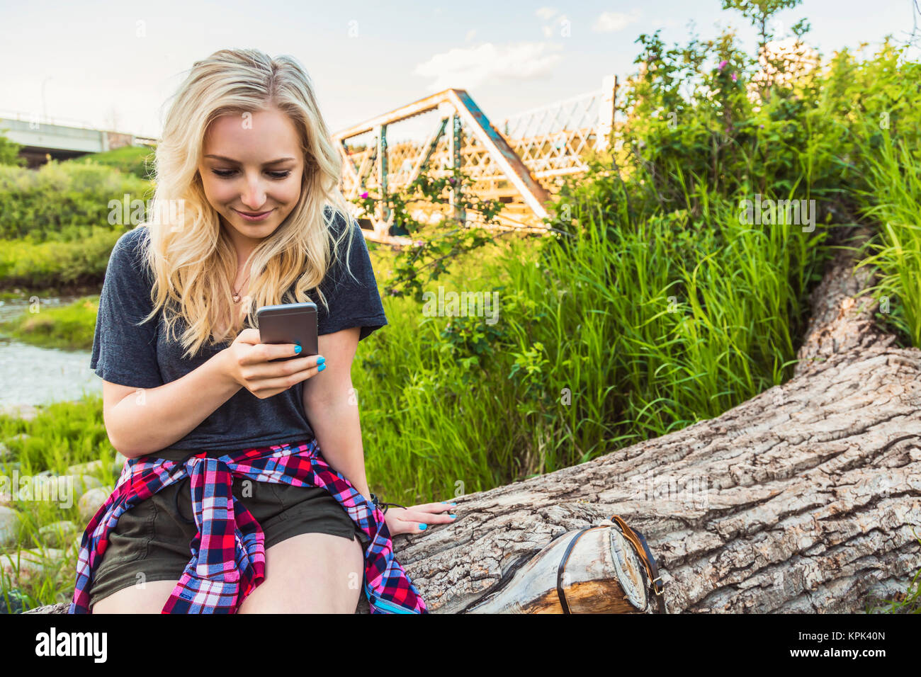 A young woman sits on log in a park using her cell phone with a river and bridge in the background; Edmonton, Alberta, Canada Stock Photo