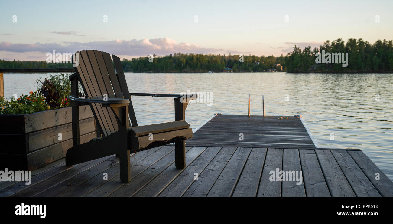 A wooden Adirondack chair sits on a dock on a tranquil lake at sunset ...