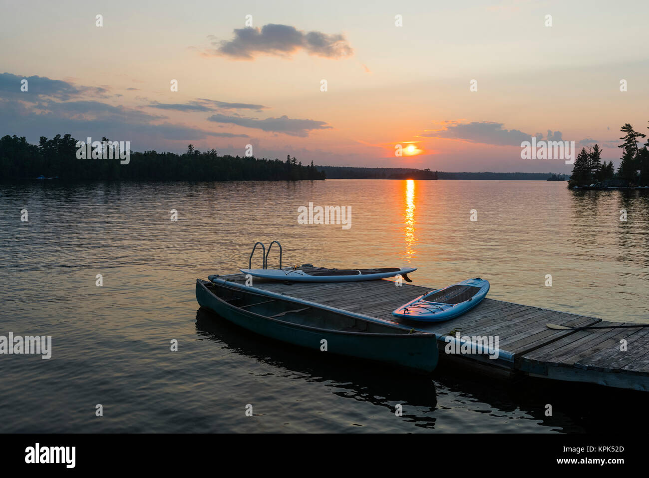The sun setting over a tranquil lake with a dock, canoe and paddle boards in the foreground; Lake of the Woods, Ontario, Canada Stock Photo