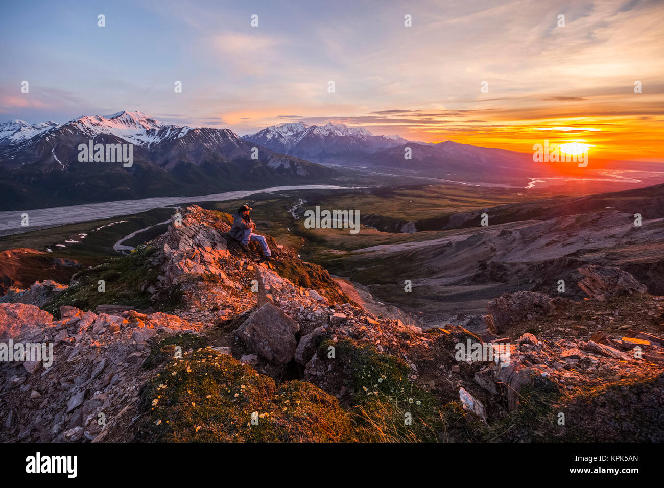 A man observes a tranquil sunset from an alpine perch high above the Delta River in the Alaska Range; Alaska, United States of America Stock Photo