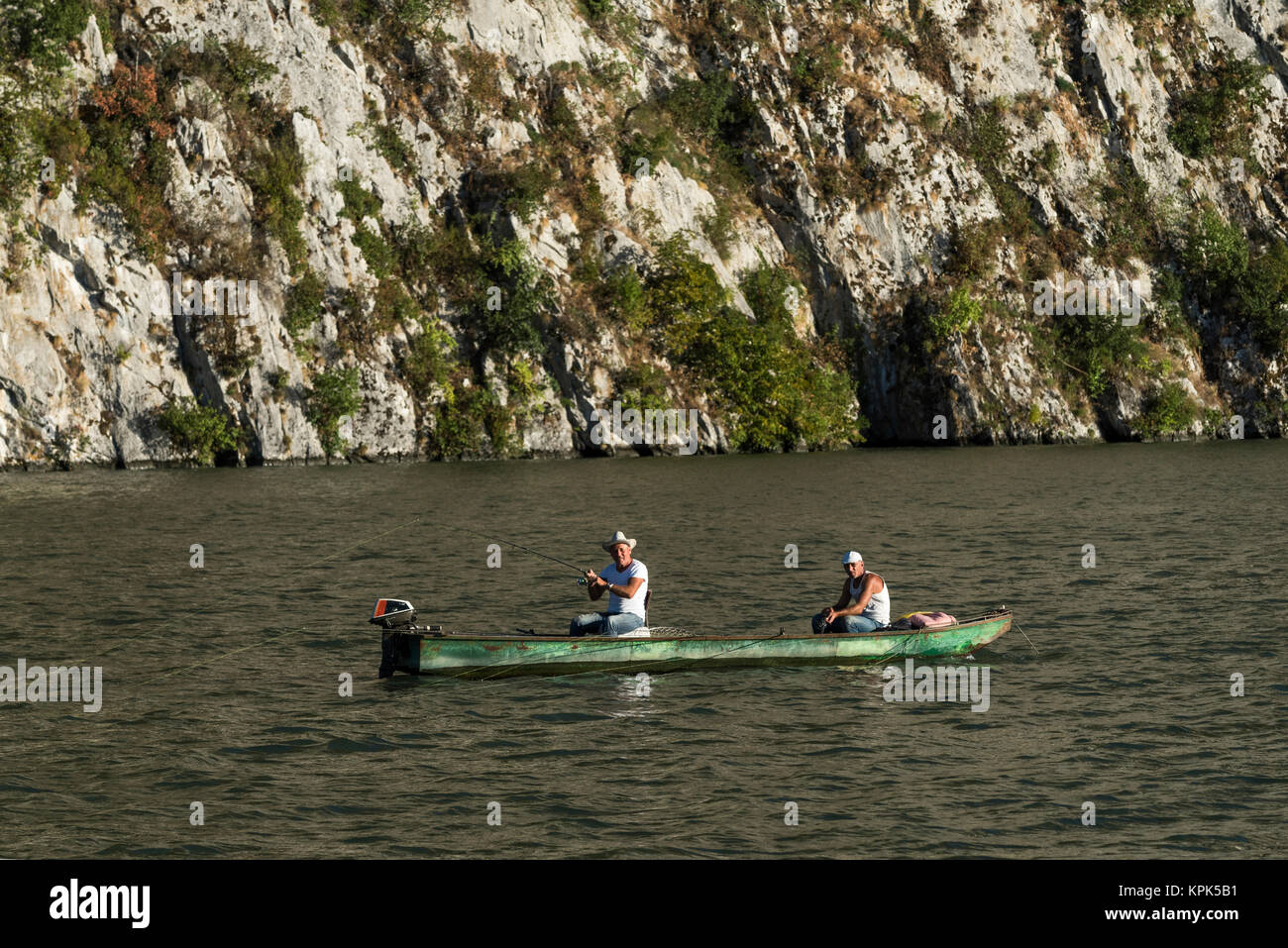 Two men sitting in a wooden boat with motor and fishing in the Danube River with a rock cliff in the background; Golubinje, Majdanpek, Serbia Stock Photo