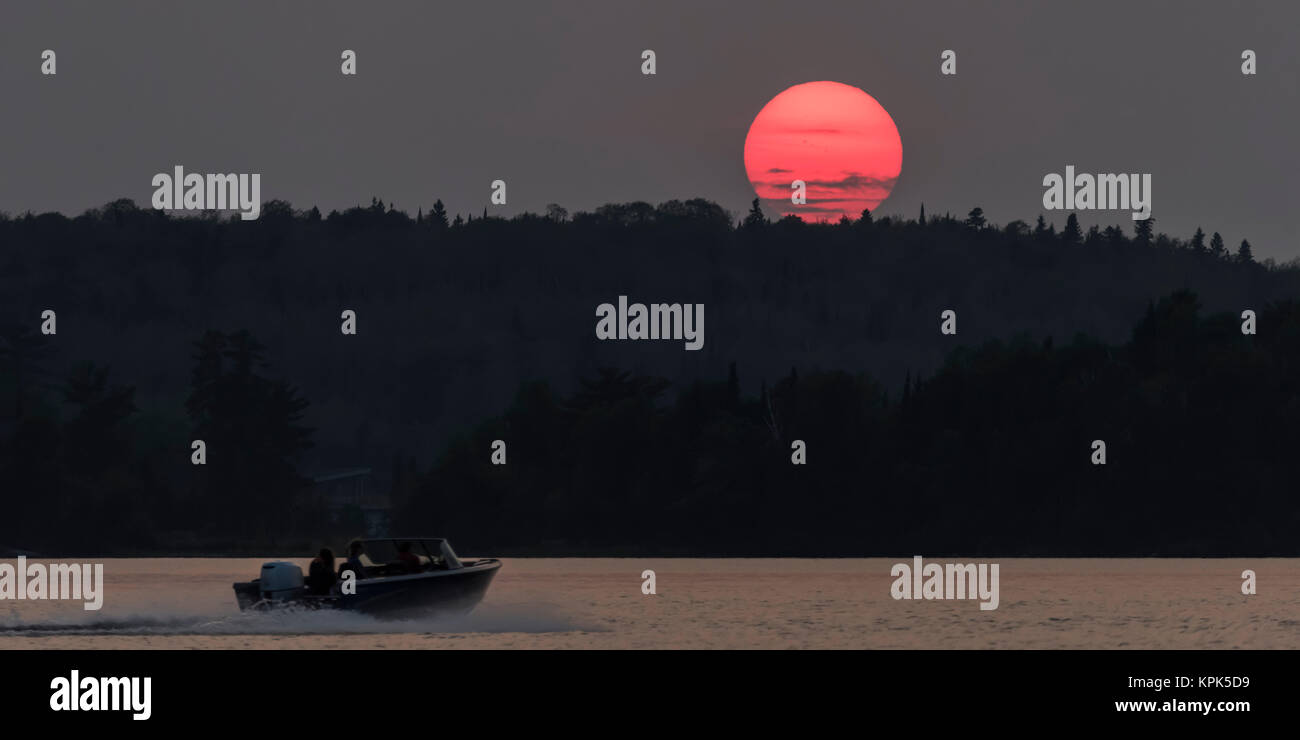 A bright pink sun sinking below a silhouetted hill and forest and a motorboat splashes on a lake; Kenora, Ontario, Canada Stock Photo