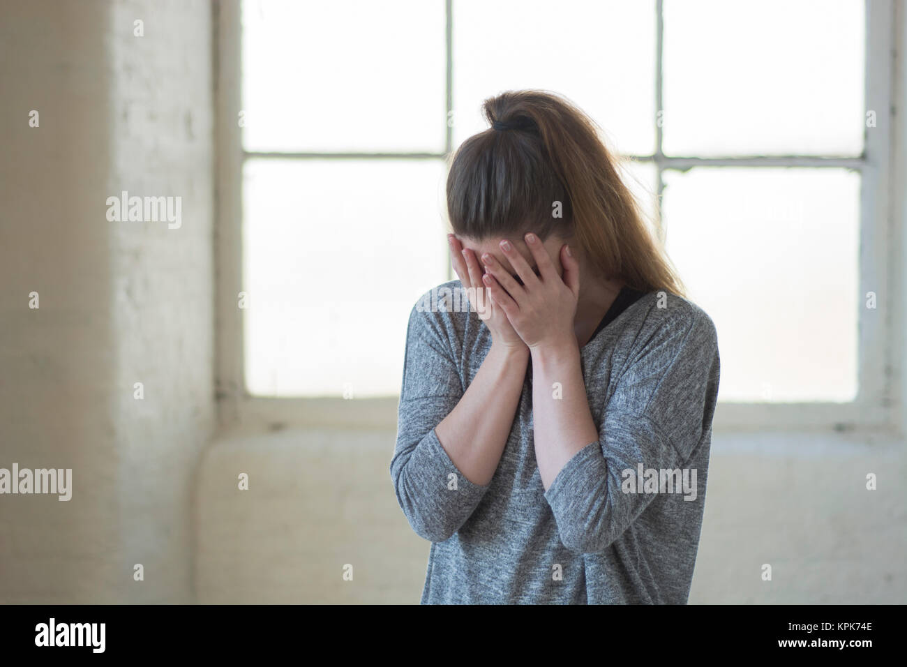 Upset young woman hiding face with hands crying by the window Stock Photo