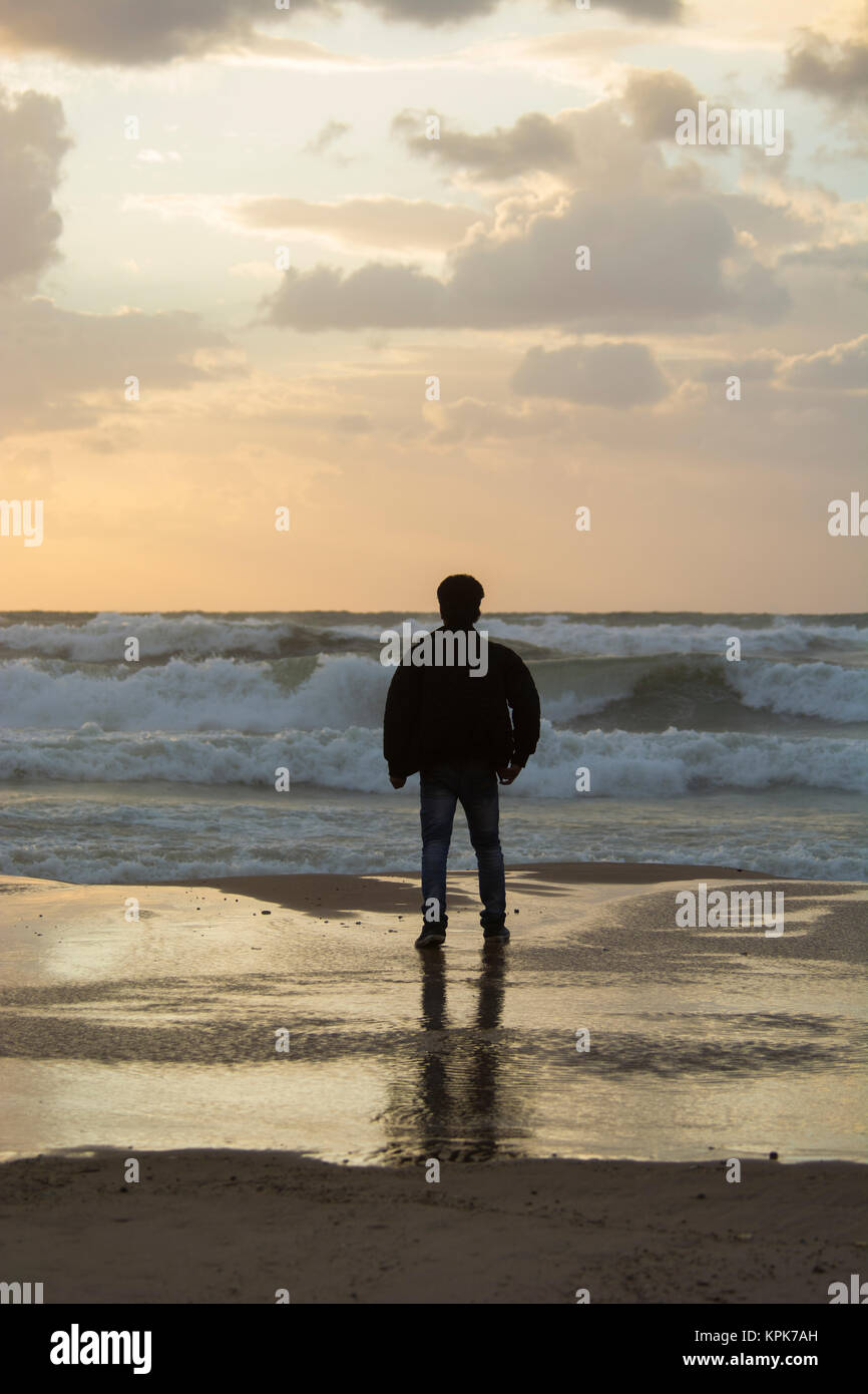 Rear view of a young silhouetted man standing on the beach at sunset ...
