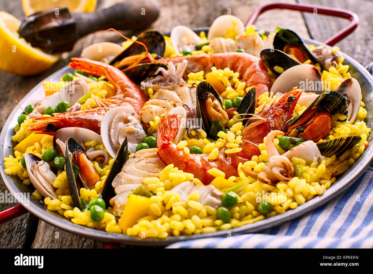 Close Up of Colorful Spanish Seafood Paella Dish with Langostina and Shellfish Served in Shallow Pan with Red Handles on Rustic Wooden Table with Fresh Lemon Garnish in Background Stock Photo