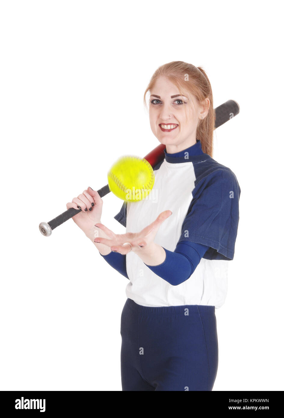 A smiling young woman standing in her blue softball uniform playing with the yellow ball holding her bat, isolated for white background Stock Photo