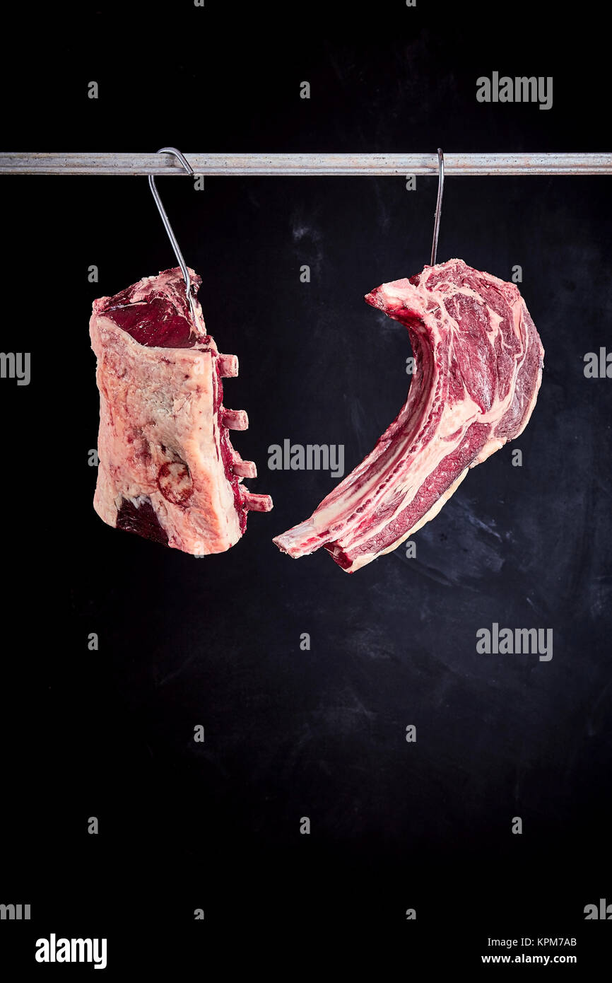 Two pieces of recently butchered raw uncooked cote de boeuf and tomahawek steak hanging from hooks on meat rack with copy space over black background Stock Photo
