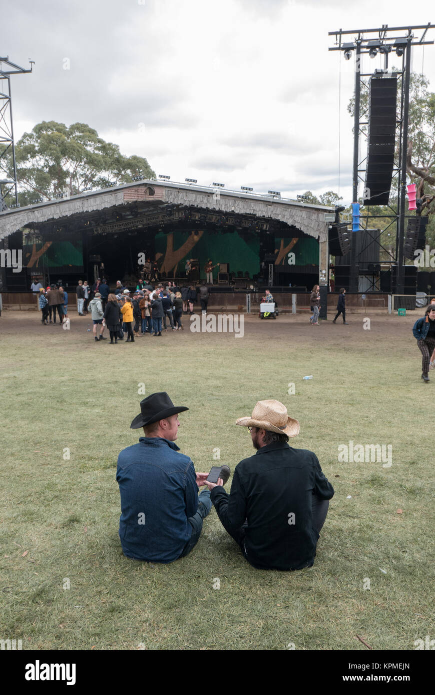 Two young Australian guys wearing black and straw cowboy hats sitting on lawn in front of music stage talking. Stock Photo